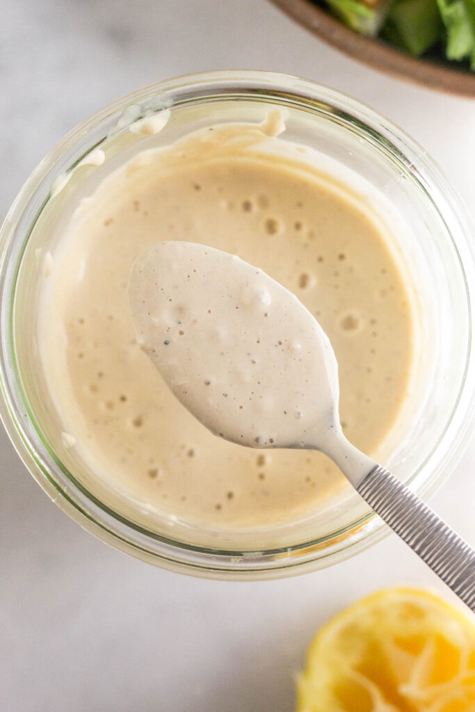 Healthy caesar dressing in a jar with a spoonful overtop the jar. Off to the side is a bowl of salad and half a juiced lemon.
