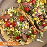 A plate of two flank steak tacos stuffed with cabbage, grilled steak, corn, tomatoes, avocado, salsa, cheese, and cilantro.