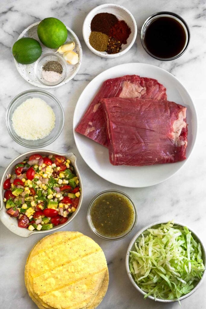 White marble counter with a bowl of spices, a bowl of coconut aminos, a plate of two pieces of raw steak, a bowl of salsa, a bowl of shredded cabbage, a stack of corn tortillas, a bowl of corn salsa, a bowl of grated cheese, and a plate of limes, garlic cloves, and small bowl of salt and pepper.