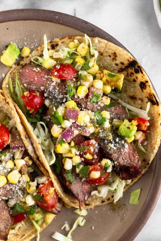 A plate steak tacos with corn tortillas stuffed with shredded cabbage, grilled steak, corn, tomatoes, avocado, cheese, and cilantro on a plate.
