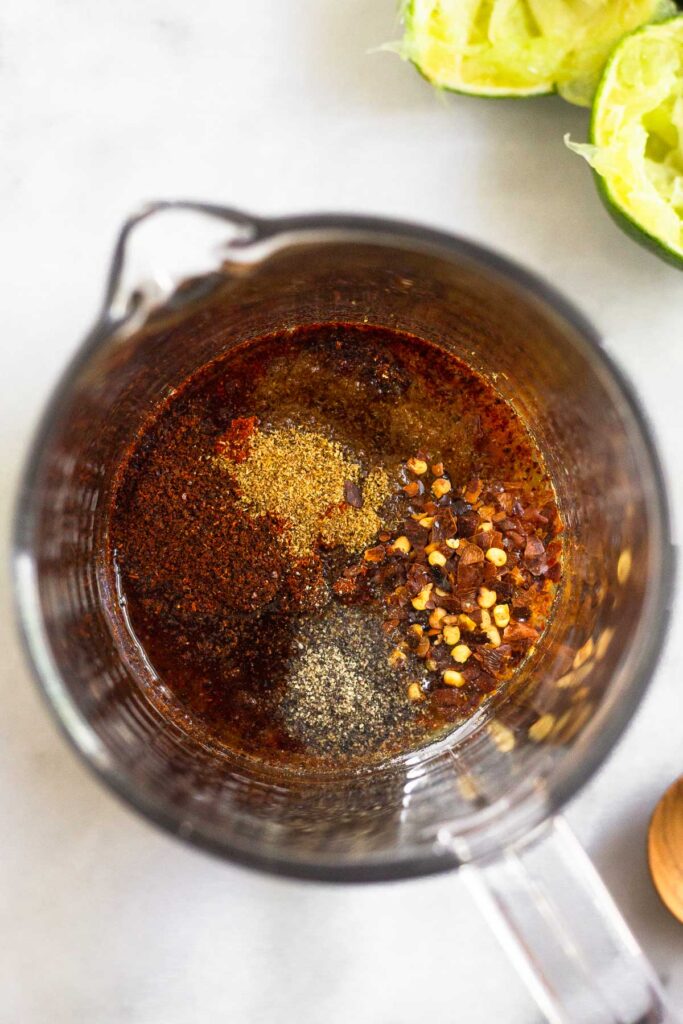Measuring cup filled with coconut aminos, lime juice, minced garlic, and spices. Next to it is 2 juiced limes.