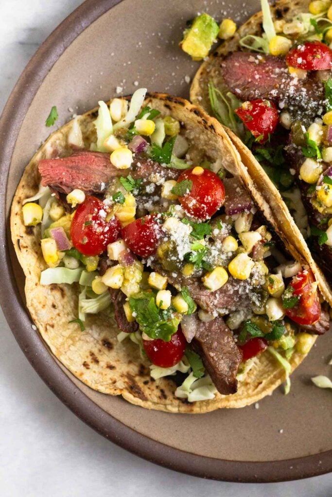 Close up of a plate with grilled steak tacos with cabbage, corn, diced tomatoes, avocado, cheese, and cilantro.