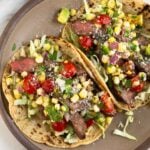 A plate of two flank steak tacos stuffed with cabbage, grilled steak, corn, tomatoes, avocado, salsa, cheese, and cilantro. Around the plate is a plate of lime wedges, a bowl of salsa, and a plate of tortillas.