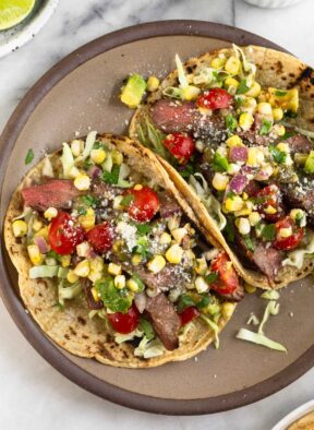 A plate of two flank steak tacos stuffed with cabbage, grilled steak, corn, tomatoes, avocado, salsa, cheese, and cilantro. Around the plate is a plate of lime wedges, a bowl of salsa, and a plate of tortillas.