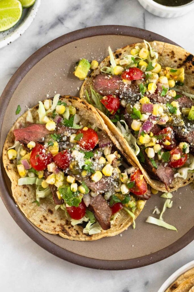 A plate of two flank steak tacos stuffed with cabbage, grilled steak, corn, tomatoes, avocado, salsa, cheese, and cilantro. Around the plate is a plate of lime wedges, a bowl of salsa, and a plate of tortillas.
