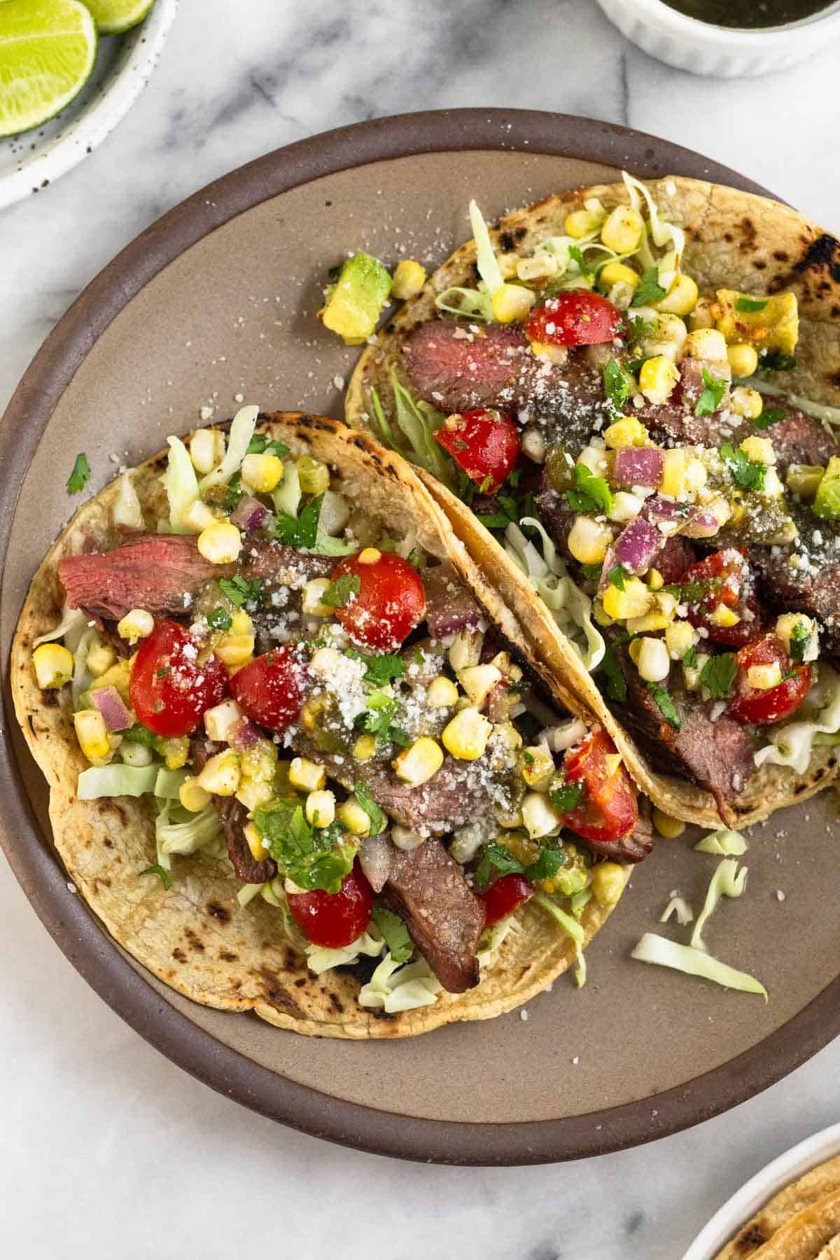 A plate of two flank steak tacos stuffed with cabbage, grilled steak, corn, tomatoes, avocado, salsa, cheese, and cilantro. Around the plate is a plate of lime wedges, a bowl of salsa, and a plate of tortillas.