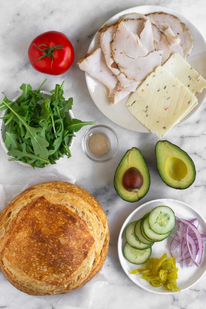 White marble counter with a plate of sliced turkey and cheese, an avocado cut in half, a plate of sliced cucumber, red onion, and banana peppers, a loaf of bread, a small bowl of mustard, a bowl of arugula, and a whole tomato.