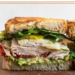 Half of a healthy sandwich on a cutting board. It has greens, banana peppers, cheese, turkey, tomato, cucumber, and avocado in it.