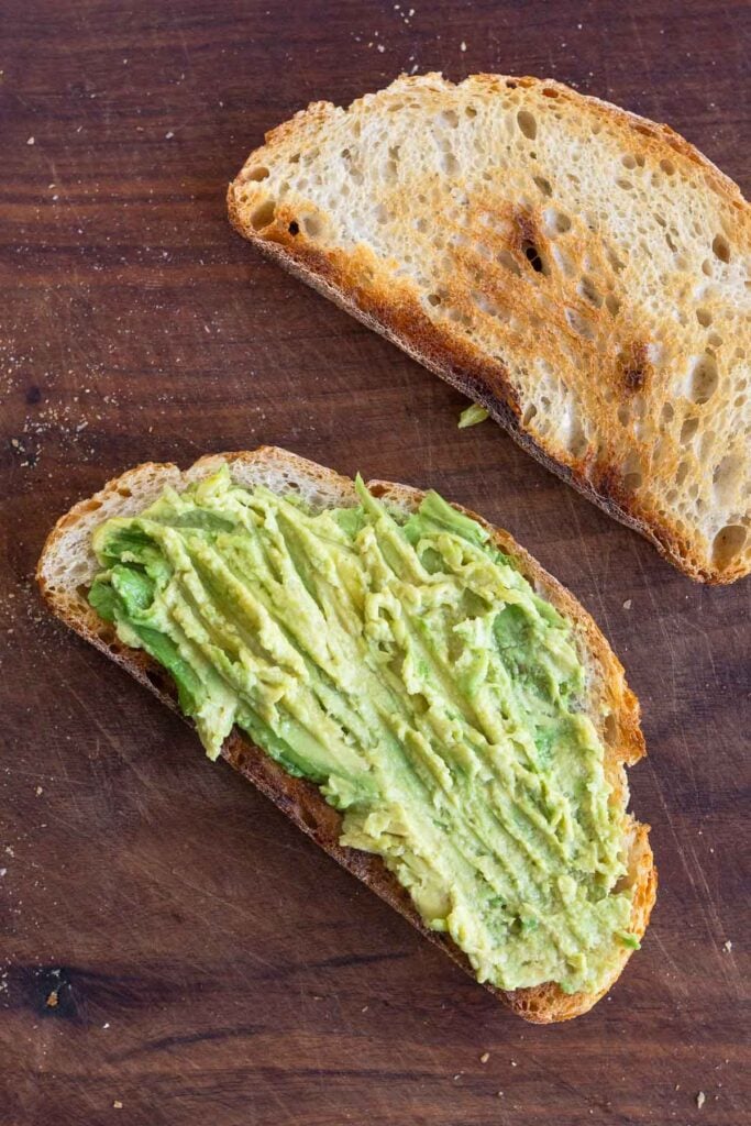 Two pieces of toasted bread on a cutting board. One piece has mashed avocado on it.