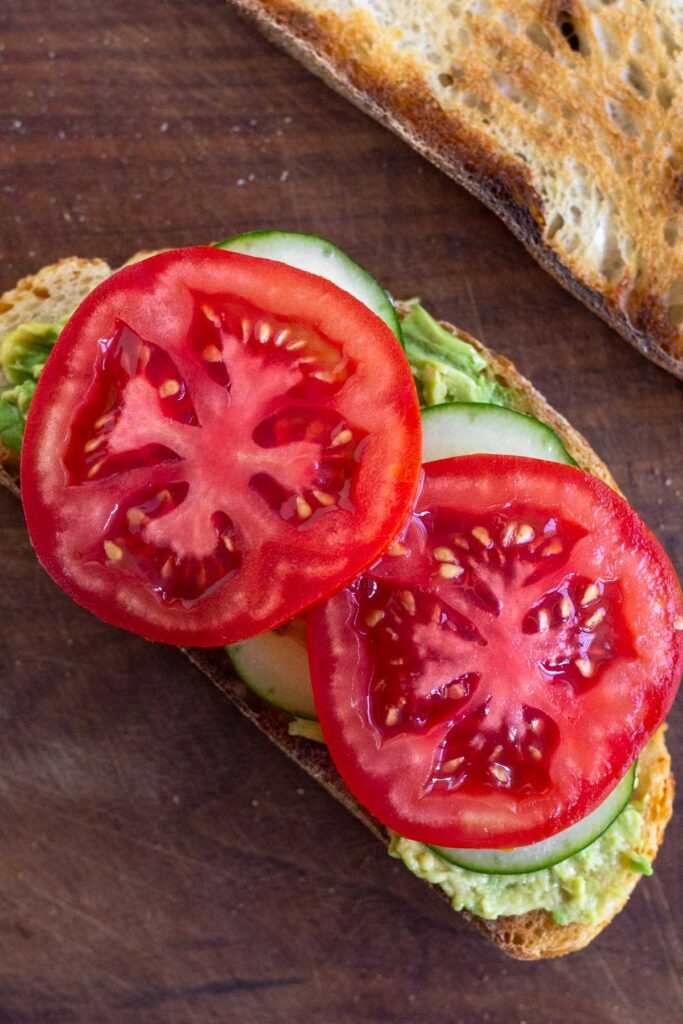 A piece of toasted bread on a cutting board topped with mashed avocado, sliced cucumber, and sliced tomato. Off to the side is another piece of toasted bread.