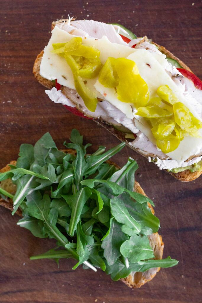 Two pieces of toasted bread on a cutting board. One piece has fresh arugula on it. The other piece has veggies, turkey, cheese, and banana peppers on it.