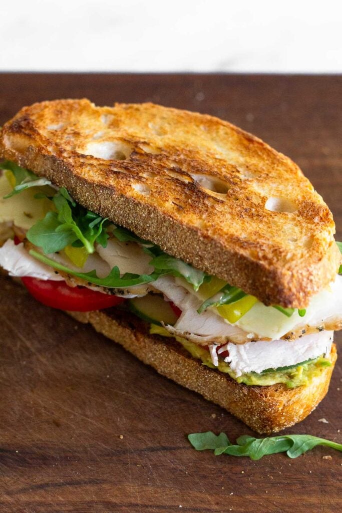 Large healthy sandwich on a cutting board with arugula, cheese, turkey, tomato, cucumber, and avocado in it.