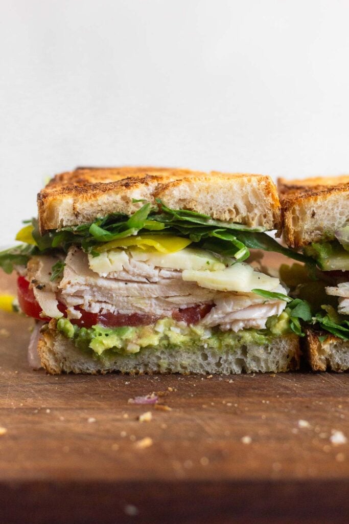 Half of a healthy sandwich on a cutting board. It has greens, banana peppers, cheese, turkey, tomato, cucumber, and avocado in it.