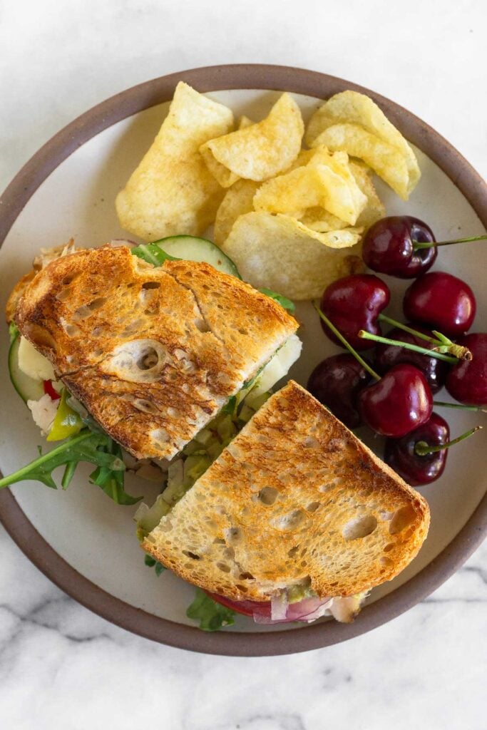 A white plate with a whole sandwich cut in half, some chips, and some cherries on it.