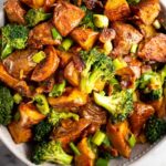 Large bowl filled with roasted potato salad with bacon and broccoli with a spoon in it. Next to the bowl is a striped towel.