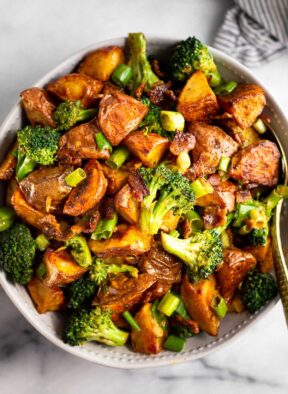 Large bowl filled with roasted potato salad with bacon and broccoli with a spoon in it. Next to the bowl is a striped towel.