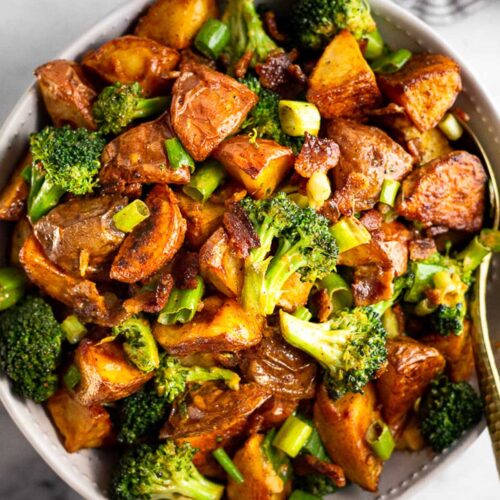 Large bowl filled with roasted potato salad with bacon and broccoli with a spoon in it. Next to the bowl is a striped towel.