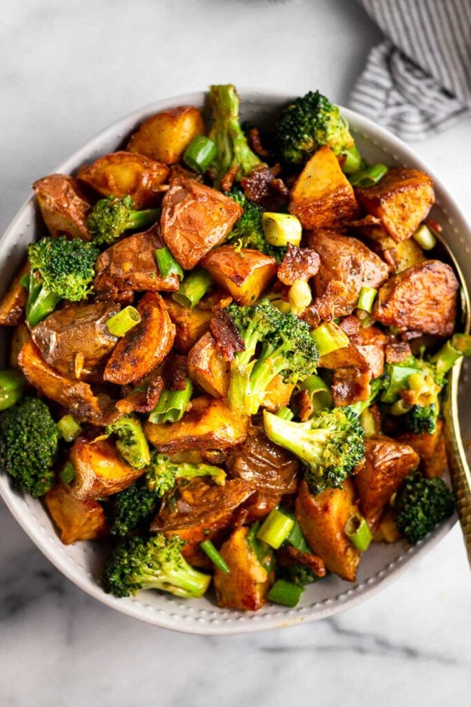 Large bowl filled with roasted potato salad with bacon and broccoli with a spoon in it. Next to the bowl is a striped towel.