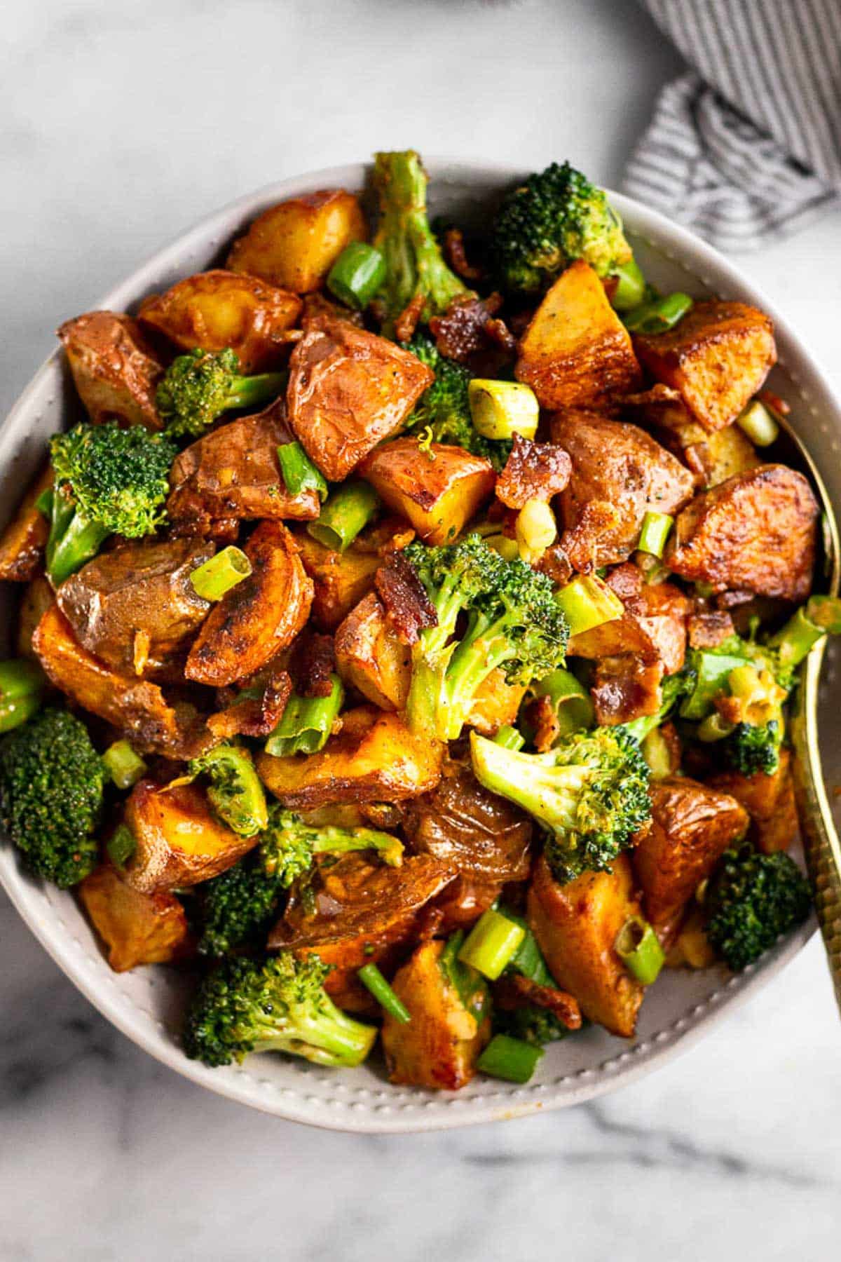 Large bowl filled with roasted potato salad with bacon and broccoli with a spoon in it. Next to the bowl is a striped towel.