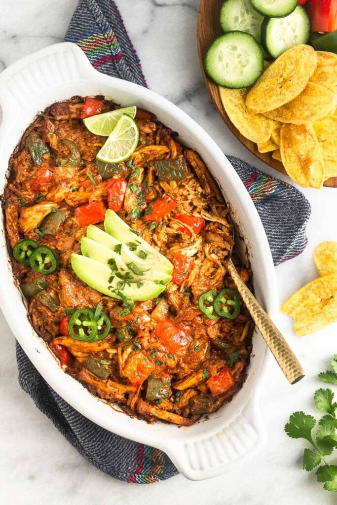 Chicken enchilada dip in a white baking dish topped with avocado, jalapeño, cilantro, and lime wedges. Next to the dish is a plate of plantains chips and veggies and a bunch of cilantro.