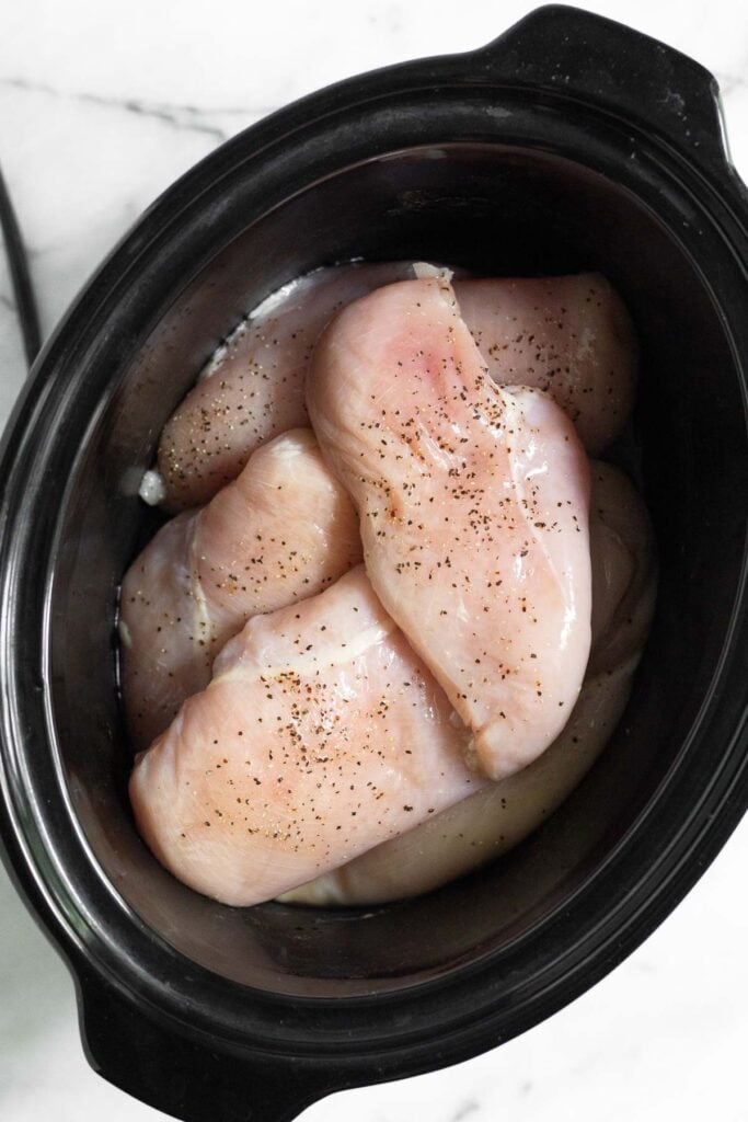 Crockpot filled with raw chicken breasts sprinkled with salt and pepper.