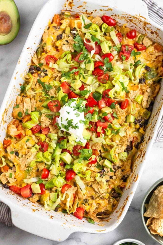 Shredded chicken taco casserole recipe in a large baking dish topped with cheese, crushed tortilla chips, shredded lettuce, diced tomatoes, sour cream, and cilantro. Around the dish is a bowl of tortilla chips and half an avocado.