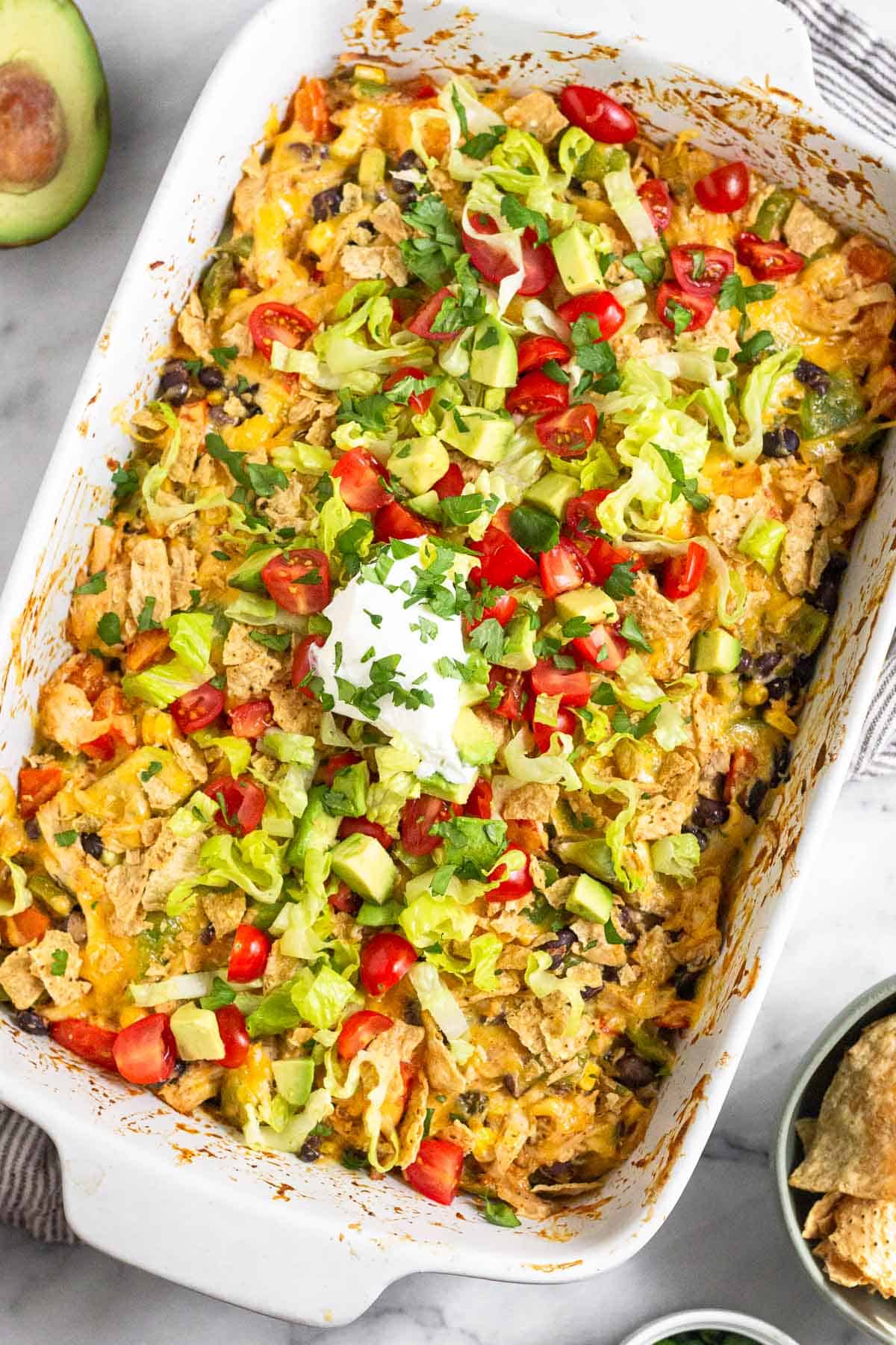 Shredded chicken taco casserole recipe in a large baking dish topped with cheese, crushed tortilla chips, shredded lettuce, diced tomatoes, sour cream, and cilantro. Around the dish is a bowl of tortilla chips and half an avocado.