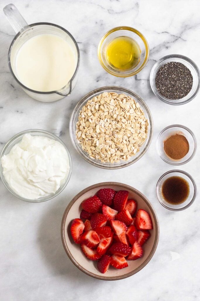 White marble counter with a small bowl of honey, a small bowl of chia seeds, a bowl of rolled oats, a small bowl of cinnamon, a small bowl of vanilla bean paste, a bowl of diced strawberries, a bowl of yogurt, and a measuring cup of milk.