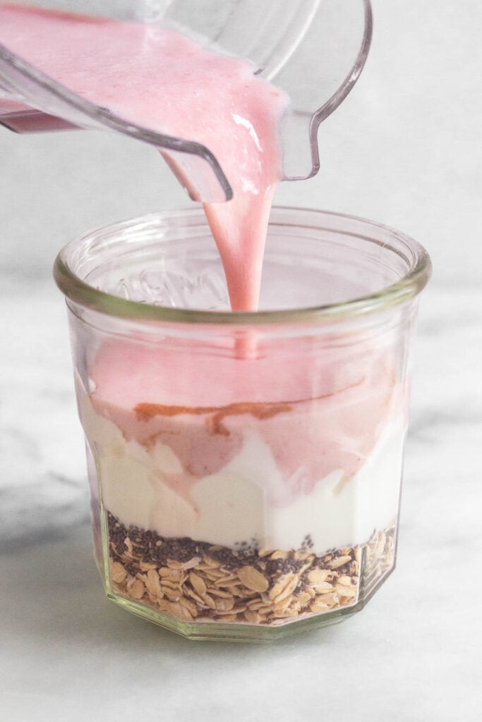 A jar layered with rolled oats, chia seeds, and greek yogurt with strawberry milk being poured on top.