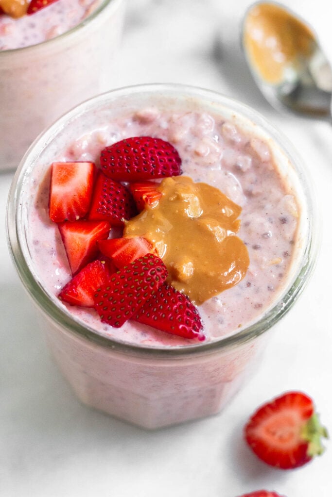 A jar of strawberries and cream overnight oats topped with diced strawberries and peanut butter. Around the jar is half a fresh strawberry, a spoon with peanut butter on it, and another jar of overnight oats.