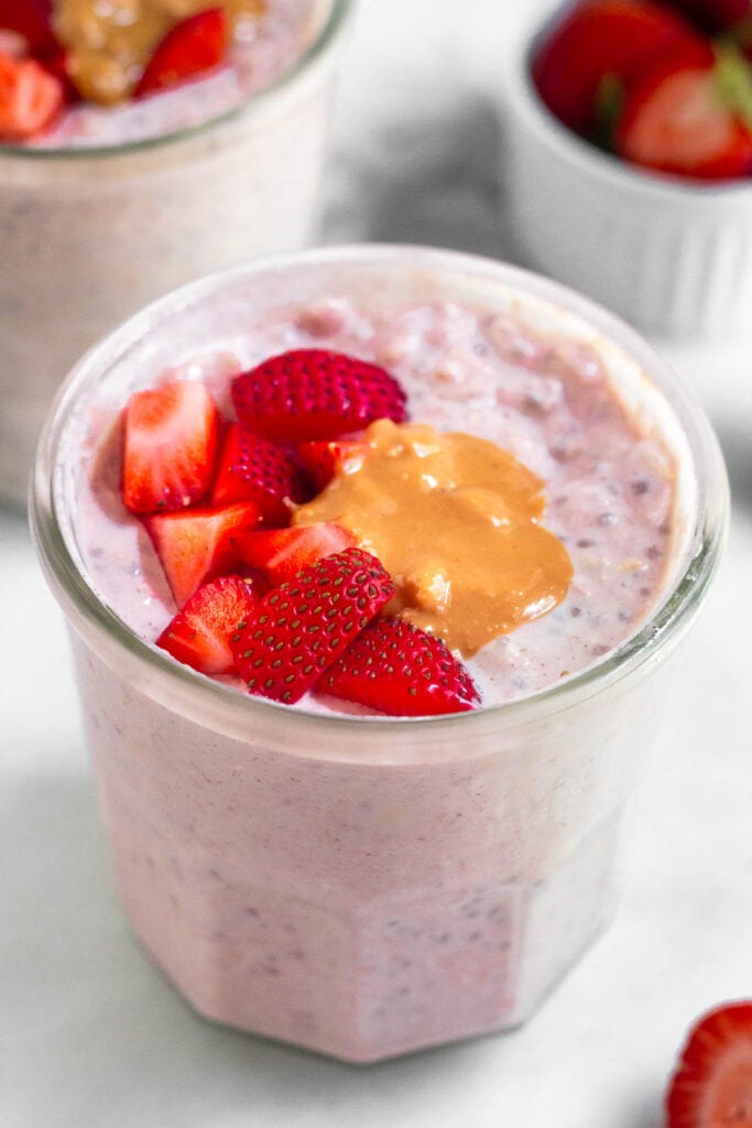 A jar of strawberry overnight oats topped with fresh diced strawberries and peanut butter. Around it is another jar of overnight oats and a small dish of strawberries.