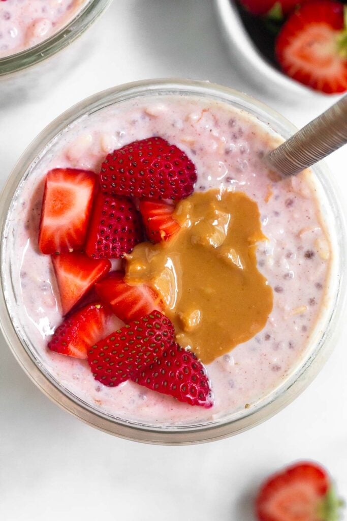 Overhead shot of a jar of strawberry overnight oats topped with diced strawberries and peanut butter with a spoon in the jar. Around the jar is a another jar of overnight oats, a bowl of diced strawberries, and half a strawberry.