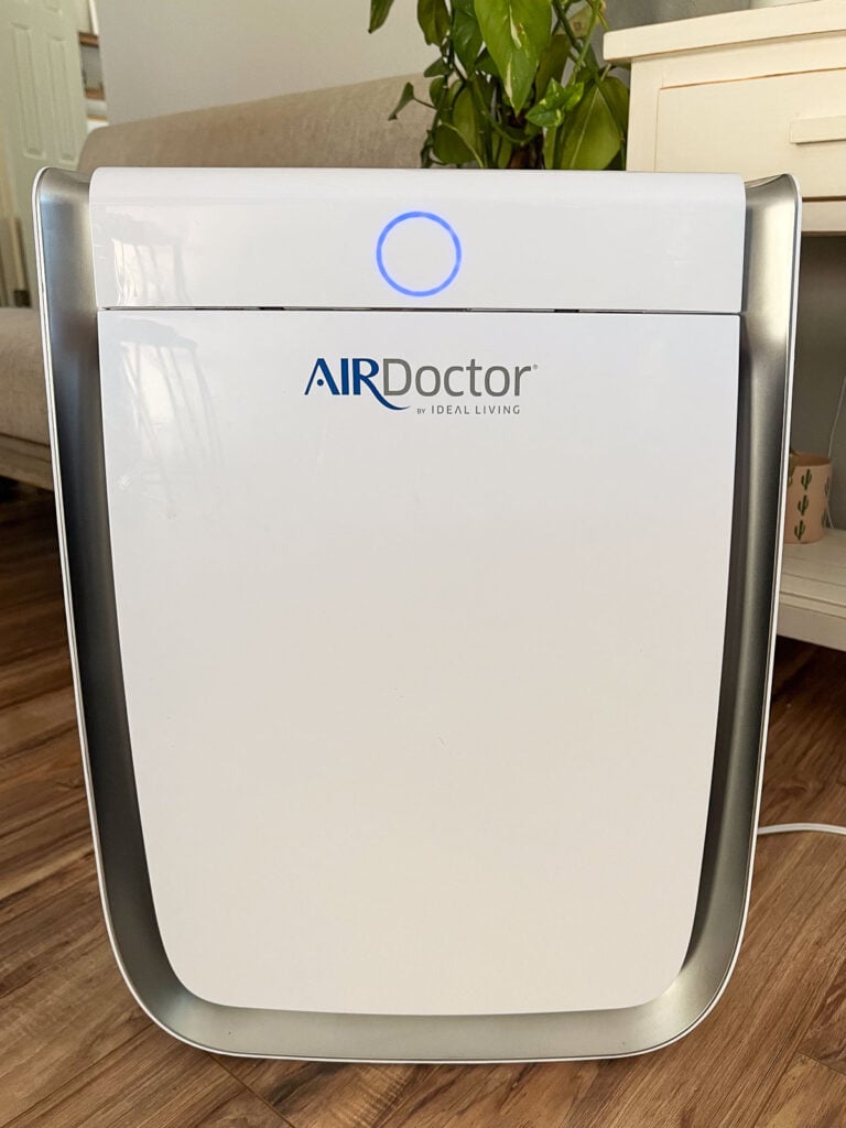 Close up of an AirDoctor air purifier in a living room. Behind it is a small couch, a plant, and a table.