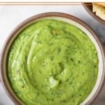 Creamy avocado sauce in a bowl. Around it is a bowl of tortilla chips, some cilantro, a lime wedge, and a striped kitchen towel.