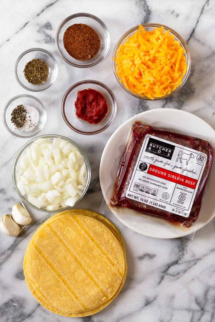Marble countertop with a bowl of shredded cheese, a package of ground beef, corn tortillas, two cloves of garlic, a bowl of diced onion, a bowl of tomato paste, a bowl of salt and pepper, a bowl of dried oregano, and a bowl of taco seasoning.