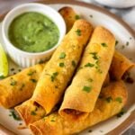 Beef taquitos stacked on top of each other on a plate. They are garnished with cilantro. Also on the plate is a ramekin of avocado sauce and a lime wedge. Behind the plate is a bowl of lime wedges and a bowl of salsa.