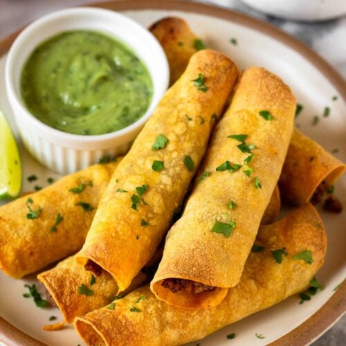 Beef taquitos stacked on top of each other on a plate. They are garnished with cilantro. Also on the plate is a ramekin of avocado sauce and a lime wedge. Behind the plate is a bowl of lime wedges and a bowl of salsa.