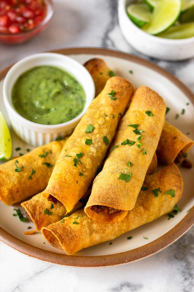 Beef taquitos stacked on top of each other on a plate. They are garnished with cilantro. Also on the plate is a ramekin of avocado sauce and a lime wedge. Behind the plate is a bowl of lime wedges and a bowl of salsa.