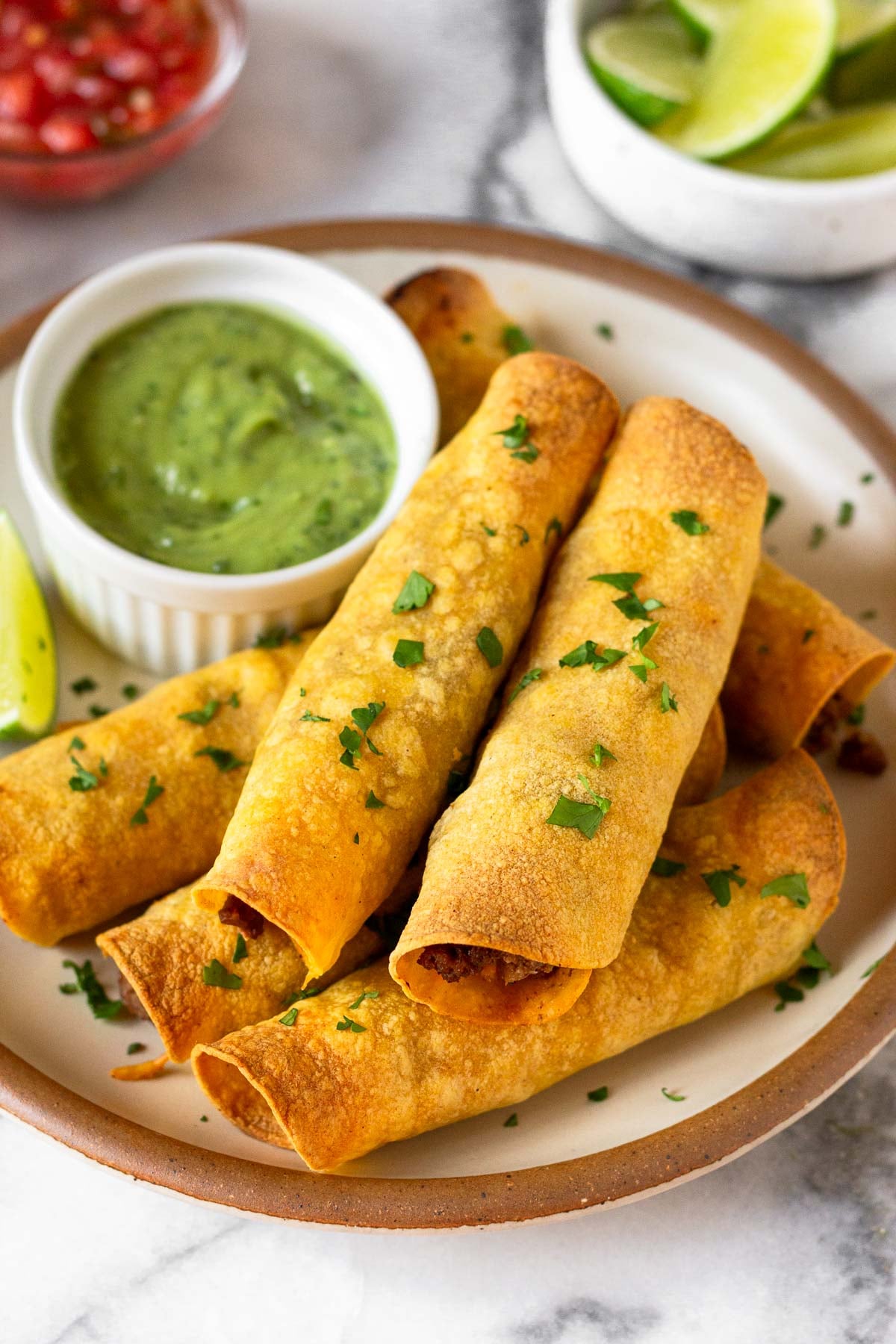 Beef taquitos stacked on top of each other on a plate. They are garnished with cilantro. Also on the plate is a ramekin of avocado sauce and a lime wedge. Behind the plate is a bowl of lime wedges and a bowl of salsa.
