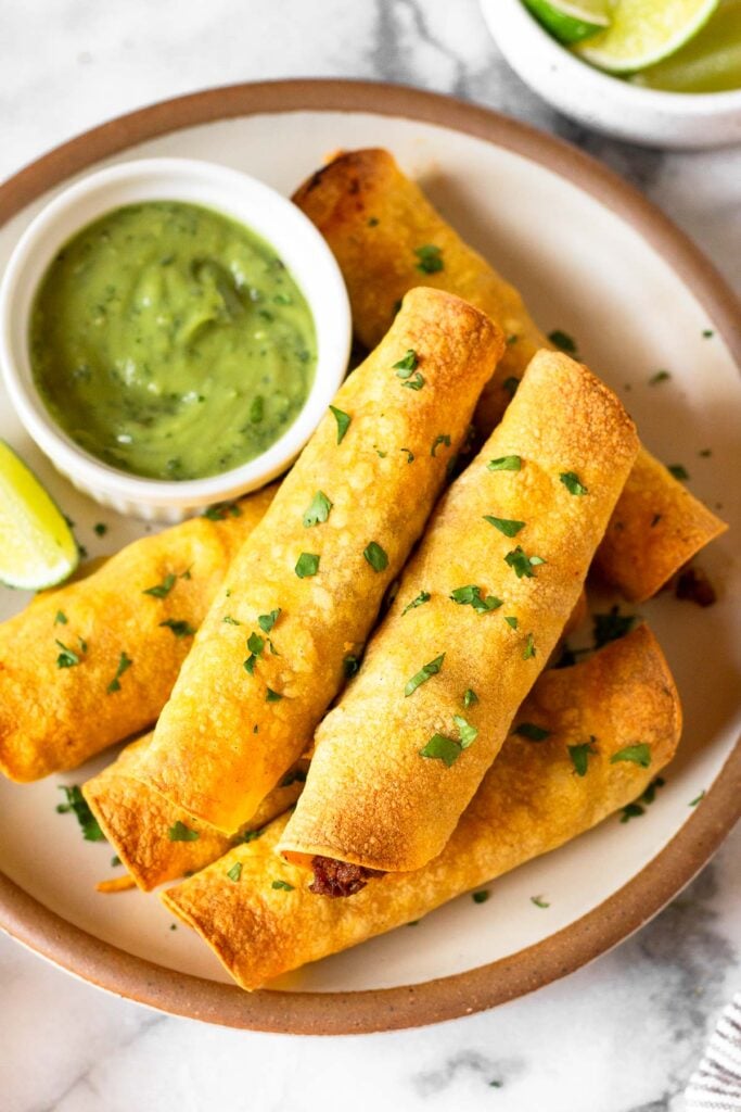 A plate of ground beef taquitos with a ramekin of avocado sauce and a lime wedge. They are garnished with chopped cilantro. Next to the plate is a bowl of lime wedges.