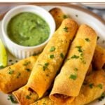 Beef taquitos stacked on top of each other on a plate. They are garnished with cilantro. Also on the plate is a ramekin of avocado sauce and a lime wedge.