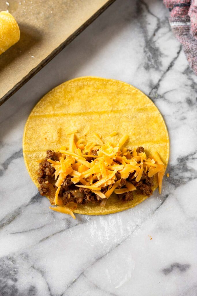Corn tortilla laying flat with half of it topped with ground beef and shredded cheese.