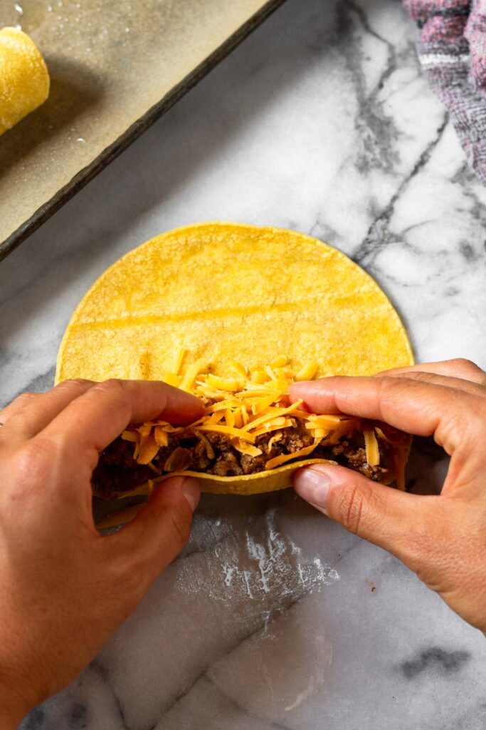Corn torilla filled with ground meat and cheese to make taquitos. There are hands on the tortilla showing it about to be rolled up.