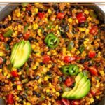 Chicken and corn skillet with black beans and veggies topped with melted cheese in a large skillet. It is garnished with sliced avocado, sliced jalapeños, and cilantro.