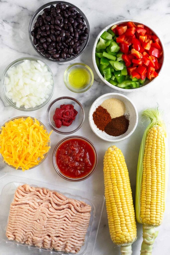 White marble counter with a bowl of black beans, a bowl of diced bell peppers, a bowl of olive oil, a bowl of spices, 2 ears of corn, a package of raw ground chicken, a bowl of salsa, a bowl of shredded cheese, a bowl of tomato paste, and a bowl of diced onions.
