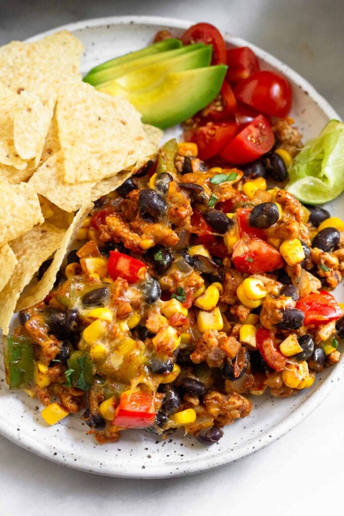A plate with mixture of chicken, black beans, and corn topped with melted cheese. Next to it are diced tomatoes, sliced avocado, and tortilla chips.