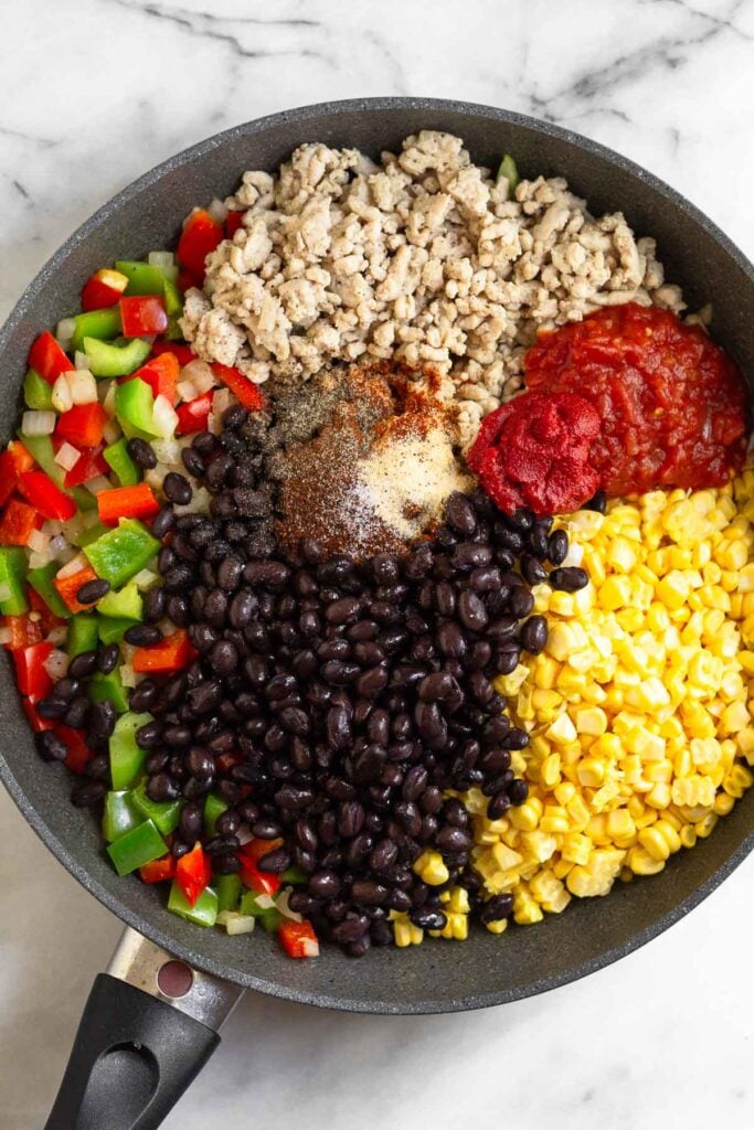Large skillet filled with sautéed onions and peppers, ground chicken, salsa, tomato paste, corn kernels, black beans, and spices before it is all mixed together.
