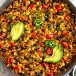 Chicken and corn skillet with veggies and black beans topped with melted cheese. It is garnished with sliced avocado, sliced jalapeños, and cilantro. Next to the skillet is half an avocado and a bowl of tortilla chips.