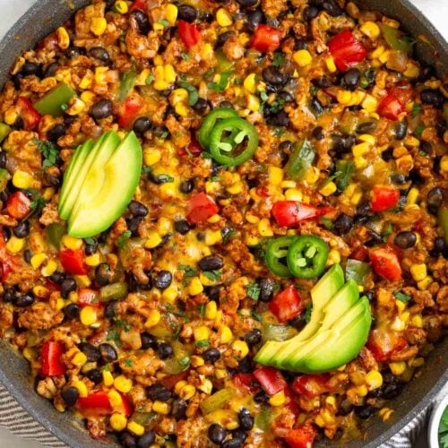Chicken and corn skillet with veggies and black beans topped with melted cheese. It is garnished with sliced avocado, sliced jalapeños, and cilantro. Next to the skillet is half an avocado and a bowl of tortilla chips.