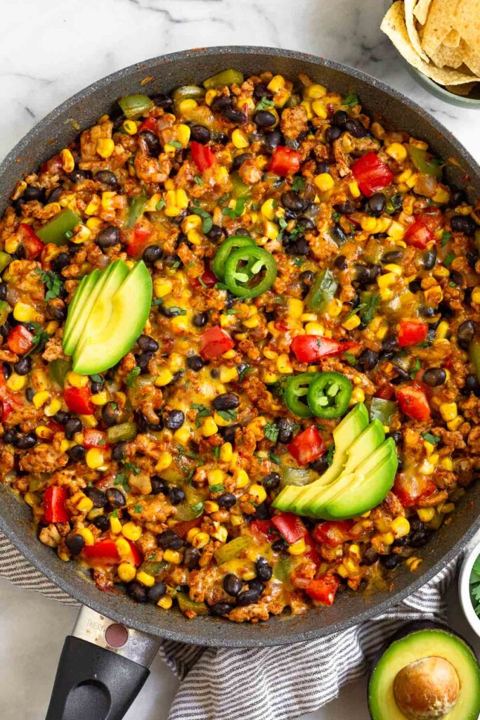 Chicken and corn skillet with veggies and black beans topped with melted cheese. It is garnished with sliced avocado, sliced jalapeños, and cilantro. Next to the skillet is half an avocado and a bowl of tortilla chips.